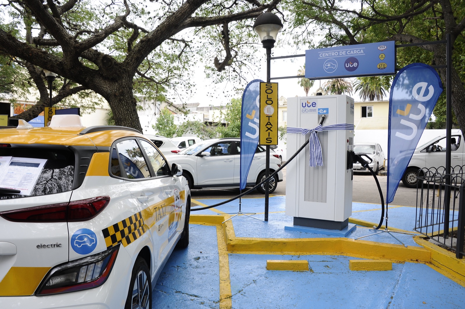 Taxi eléctrico cargando en un Centro de Carga de UTE, con banderas de Movilidad Eléctrica