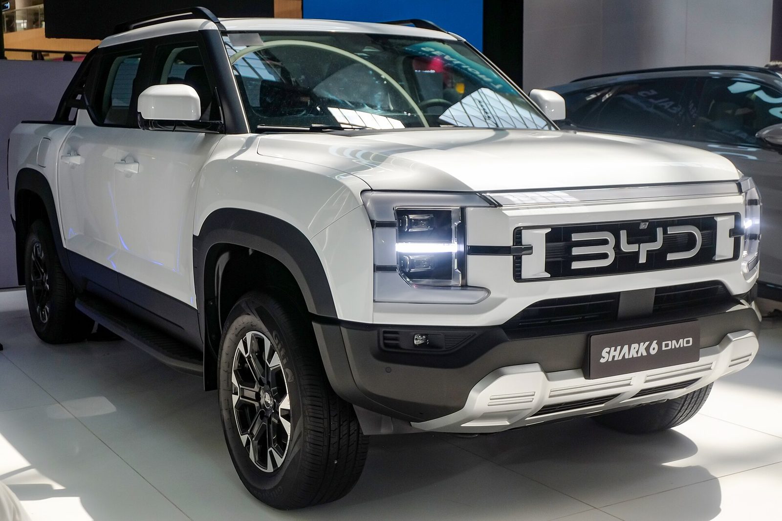 BYD Shark 6 DMO blanca de frente en un salón de exhibición, con el logo BYD en la parrilla y la identificación del modelo en la matrícula